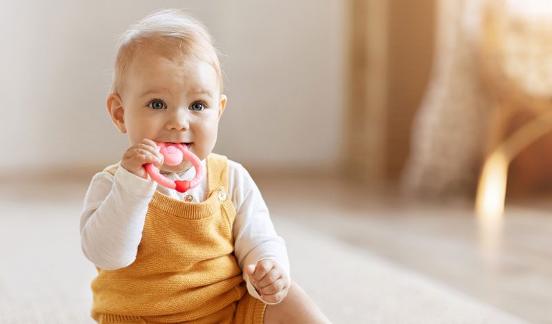 Baby sitzt auf dem Teppich und hält einen Beißring im Mund